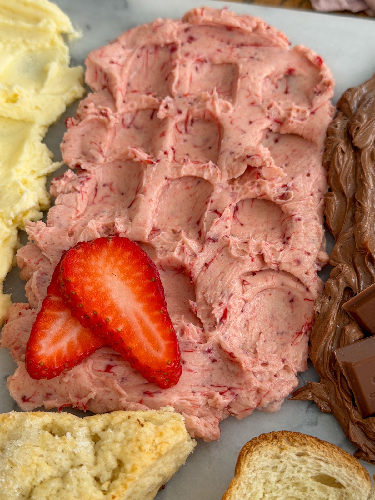 Close up of strawberry compound butter spread on a marble board with fresh sliced strawberry as garnish and small scone pieces alongside.