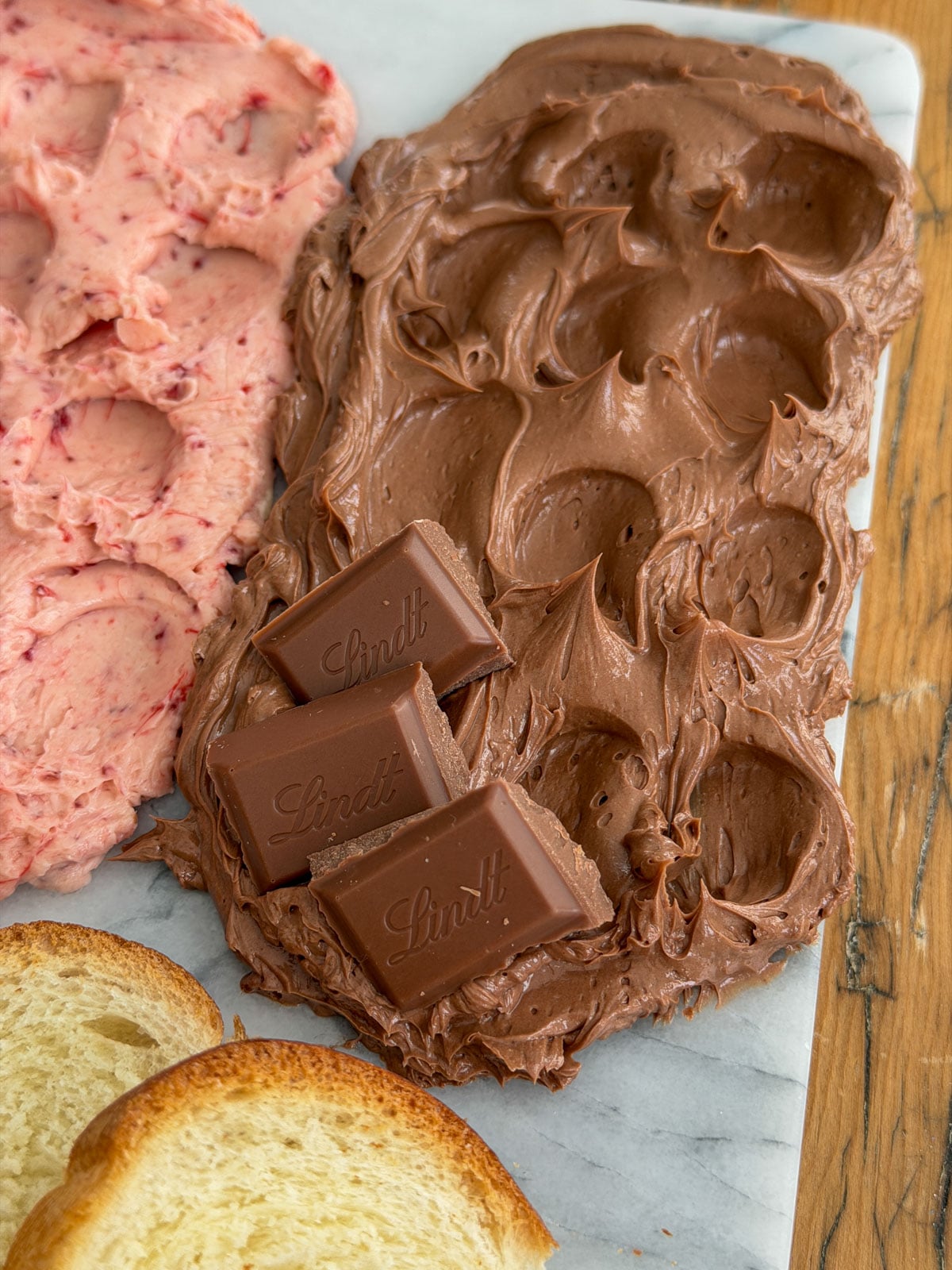 Close up of chocolate compound butter spread on a marble board with Lindt chocolate pieces as garnish and sliced brioche alongside.