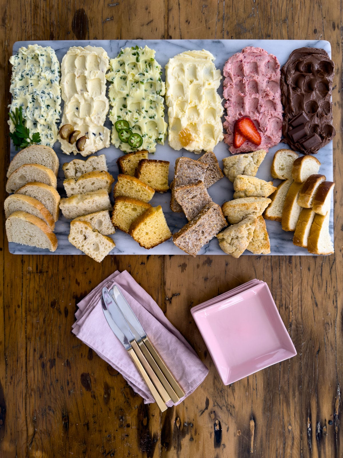 Overhead view of a complete compound butter board with six compound butters and paired breads on a marble board, styled with pink plates, gold butter knives, linen napkins, and wine glasses on a wood table.