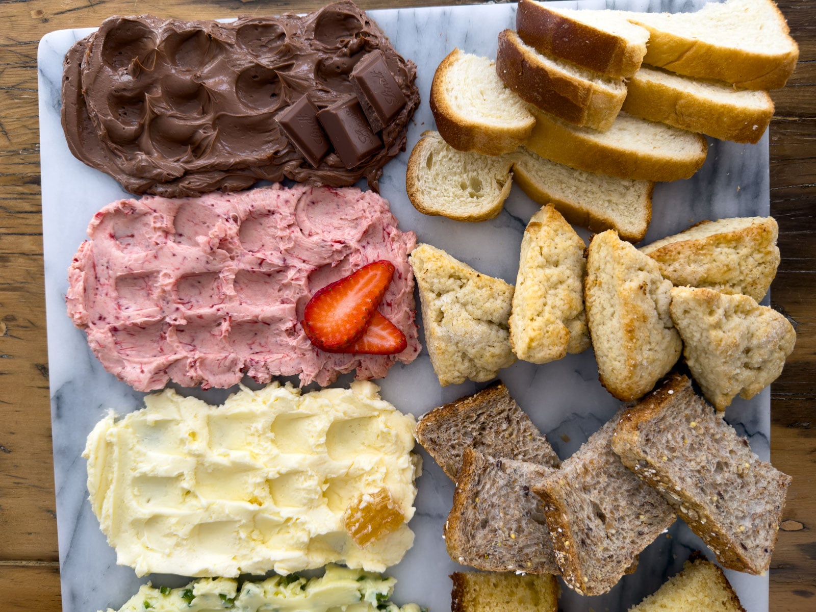 Close up of strawberry compound butter and chocolate compound butter on a marble board with scones and sliced brioche alongside