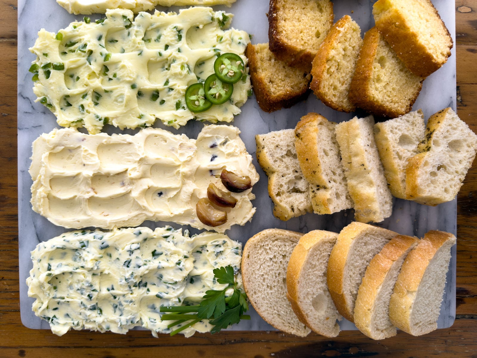 Overhead view of three savory compound butters on a marble board including jalape&ntilde;o honey butter, roasted garlic butter, and garlic herb butter, each paired with its matching bread of cornbread slices, focaccia, and sourdough.