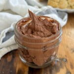 Whipped chocolate butter in a small glass jar with a swirled peak, with scones on a white plate in the background.