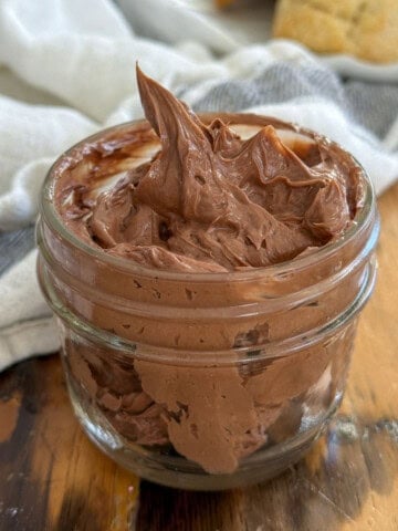 Whipped chocolate butter in a small glass jar with a swirled peak, with scones on a white plate in the background.
