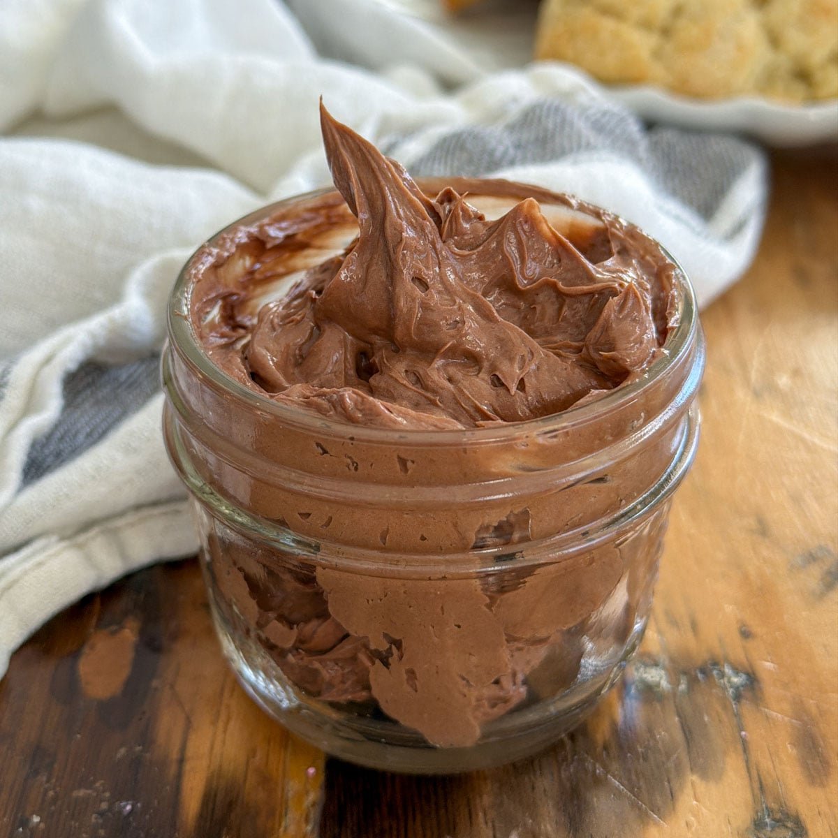 Whipped chocolate butter in a small glass jar with a swirled peak, with scones on a white plate in the background.