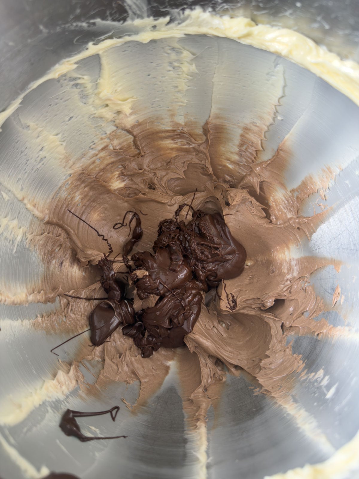 Melted chocolate added to whipped butter mixture in a stand mixer bowl showing the two components beginning to combine.