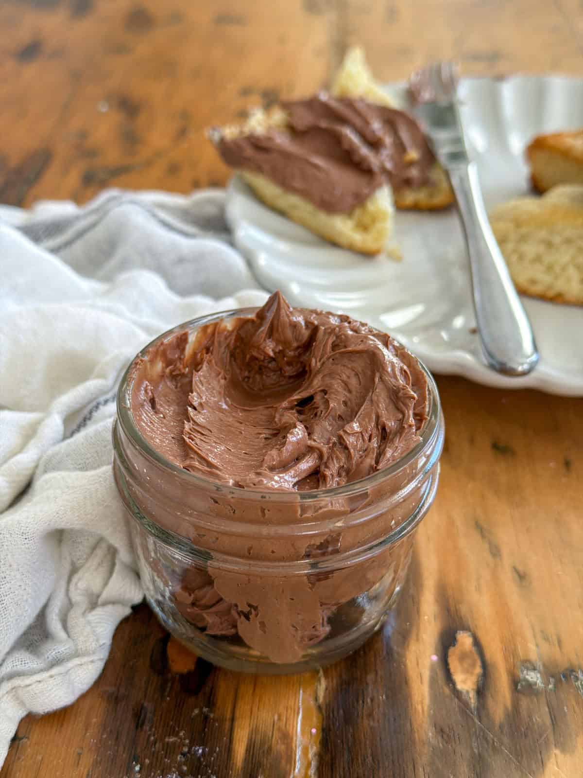 Chocolate butter spread thickly on a split scone on a white scalloped plate with a butter knife and jar of chocolate butter in the background.