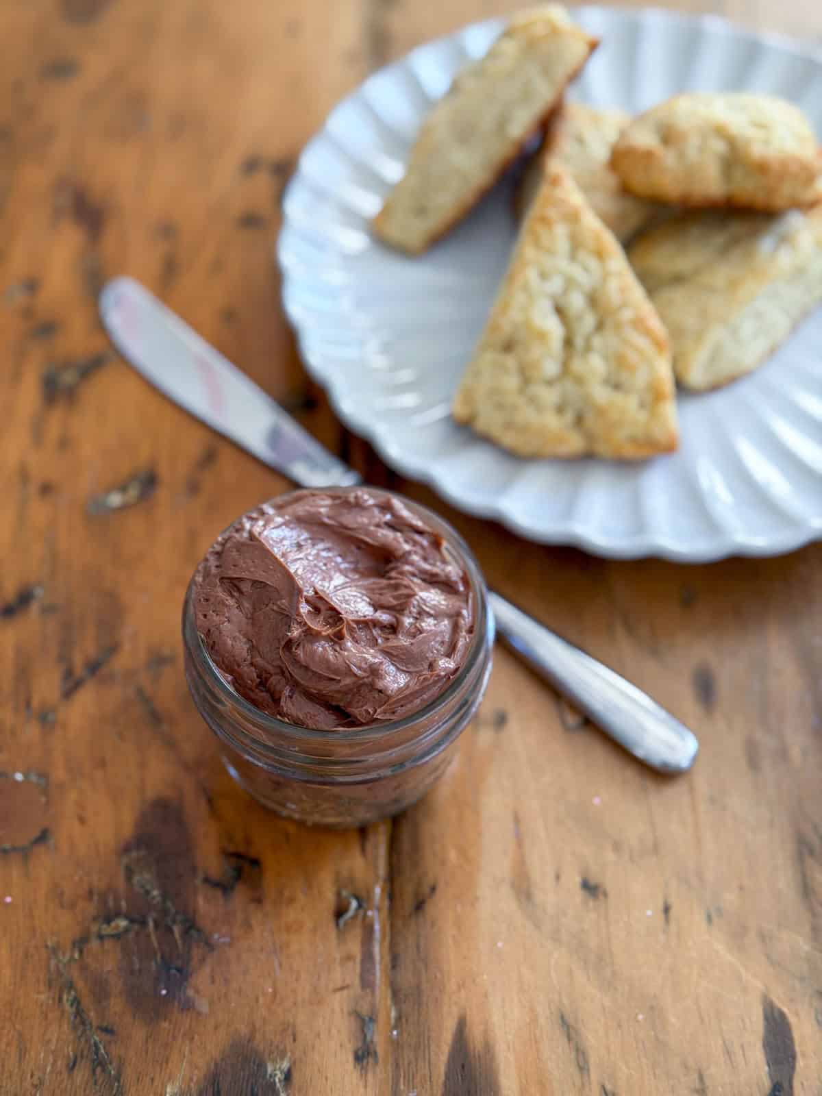 Glass jar of whipped chocolate butter on a wooden table with a white scalloped plate of scones and a silver butter knife alongside.