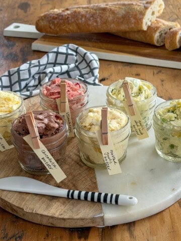 Six compound butter jars with handwritten clothespin labels showing all six flavors including whipped honey butter, chocolate butter, strawberry butter, roasted garlic butter, garlic herb butter, and jalapeno honey butter arranged on a round marble and wood board with a sliced baguette on a rectangular board in the background on a rustic wooden table.