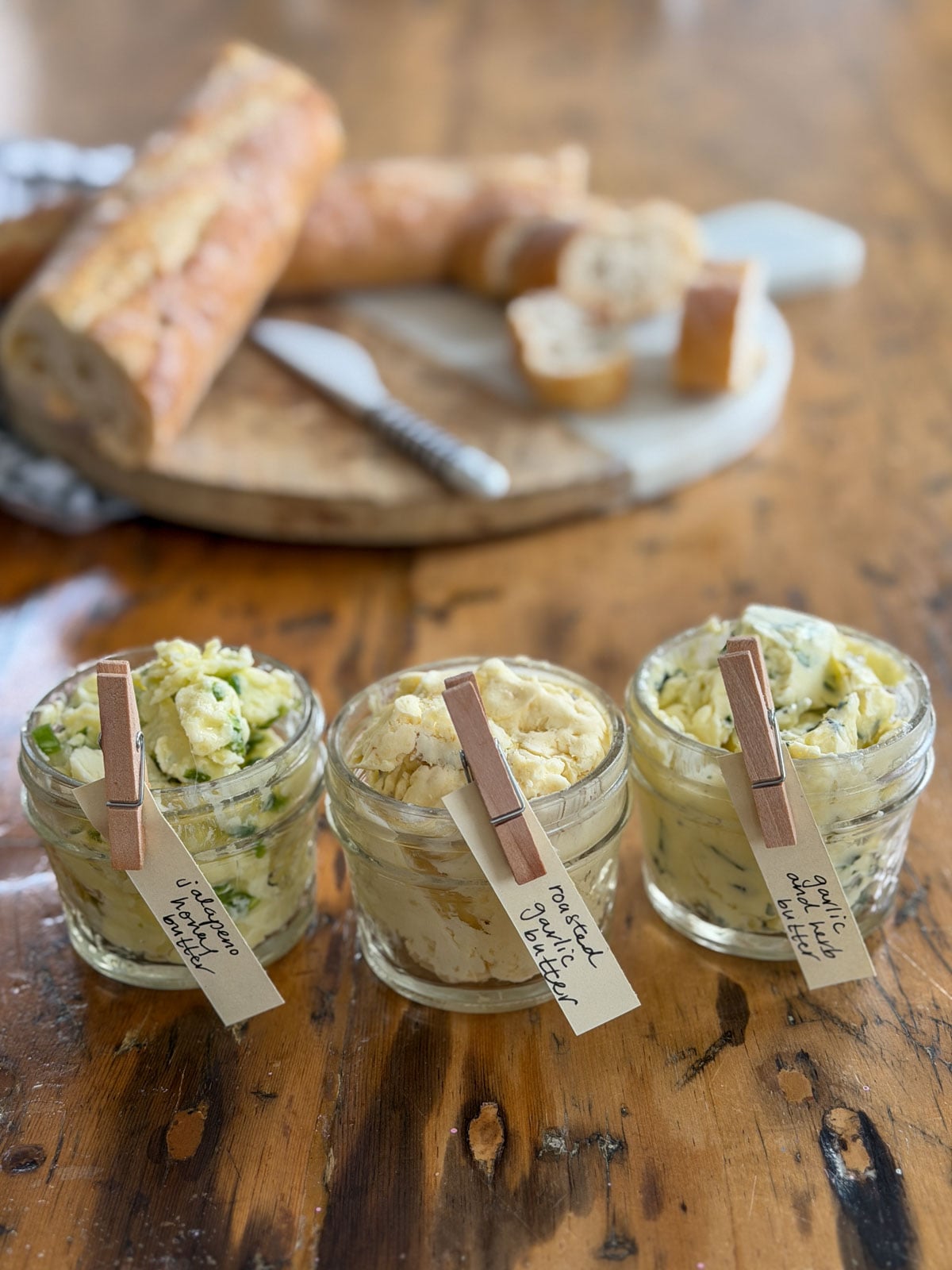 Three savory compound butter jars labeled jalapeno honey butter, roasted garlic butter, and garlic herb butter with handwritten clothespin labels on a wooden table with a baguette and bread on a marble board in the background.