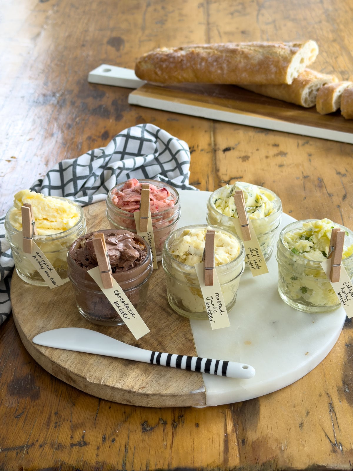 Six homemade compound butters in labeled mason jars arranged on a round marble and wood board with a sliced baguette in the background, showing how to store compound butters ahead of time