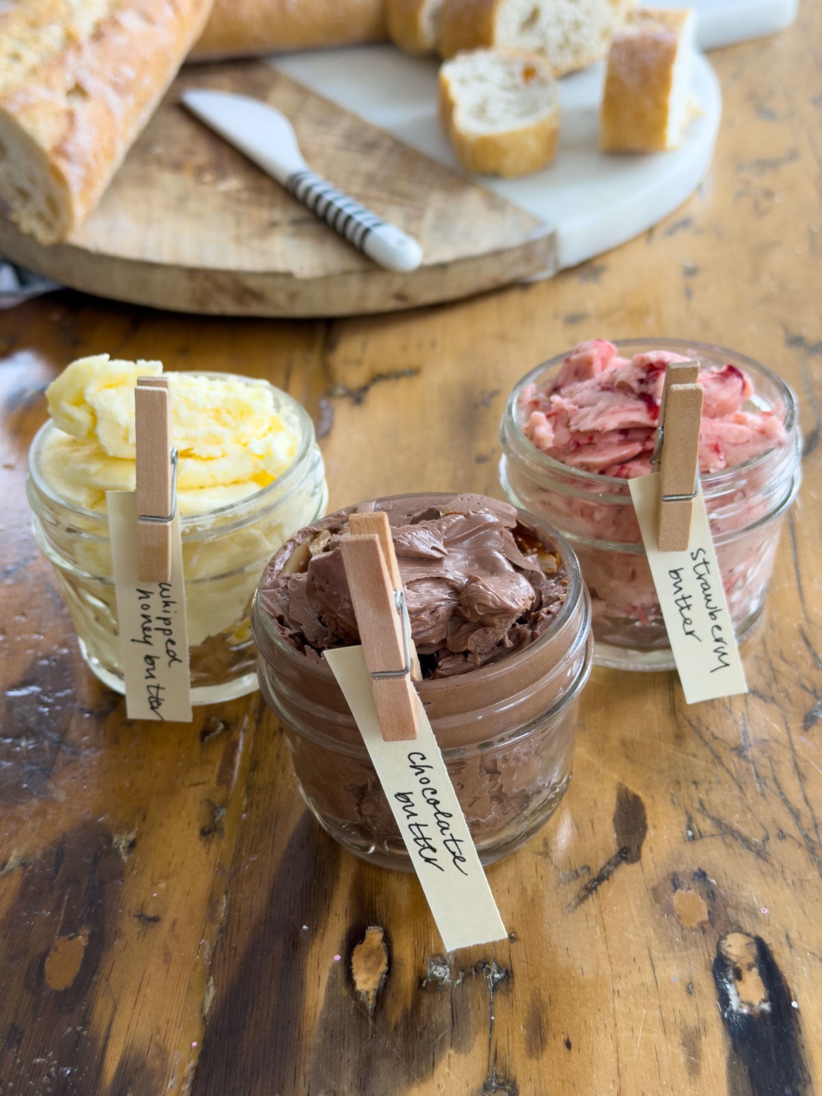 Three sweet compound butter jars labeled whipped honey butter, chocolate butter, and strawberry butter with handwritten clothespin labels on a wooden table with baguette and bread in the background.