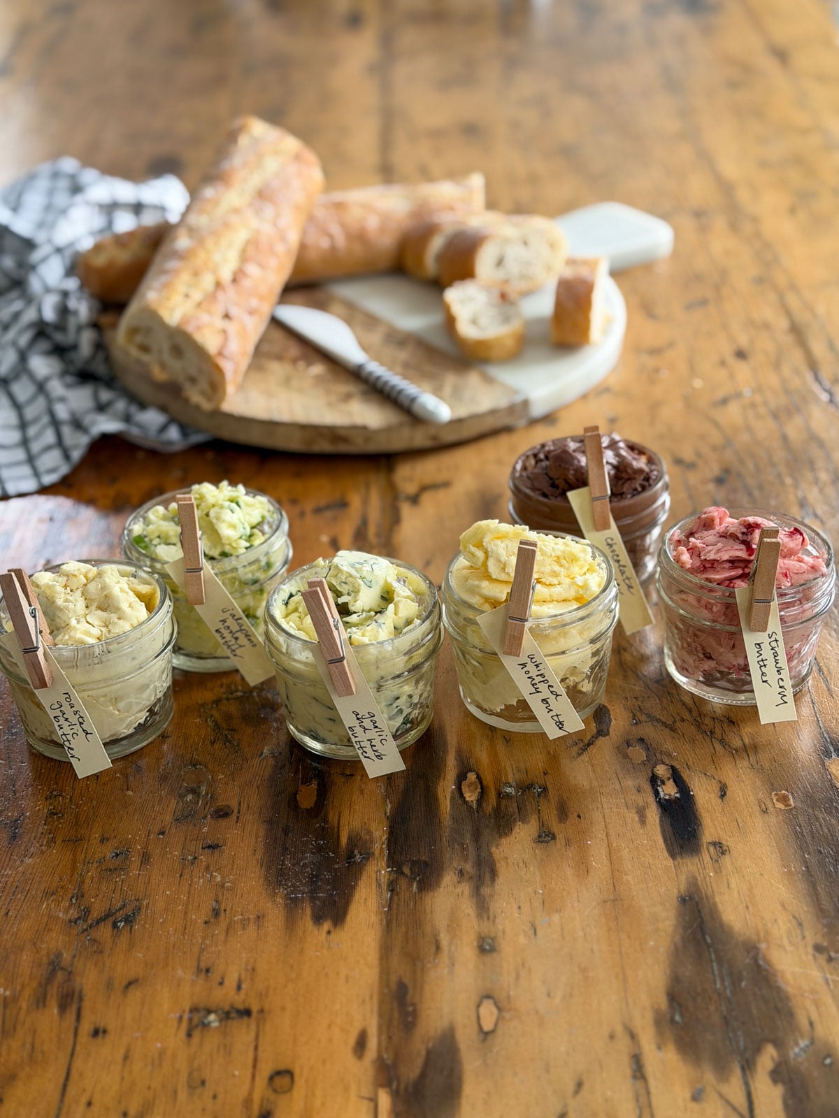 Six compound butter jars with handwritten clothespin labels showing all six flavors of homemade compound butter arranged with a round bread board in the background on a wooden table with a striped linen napkin.