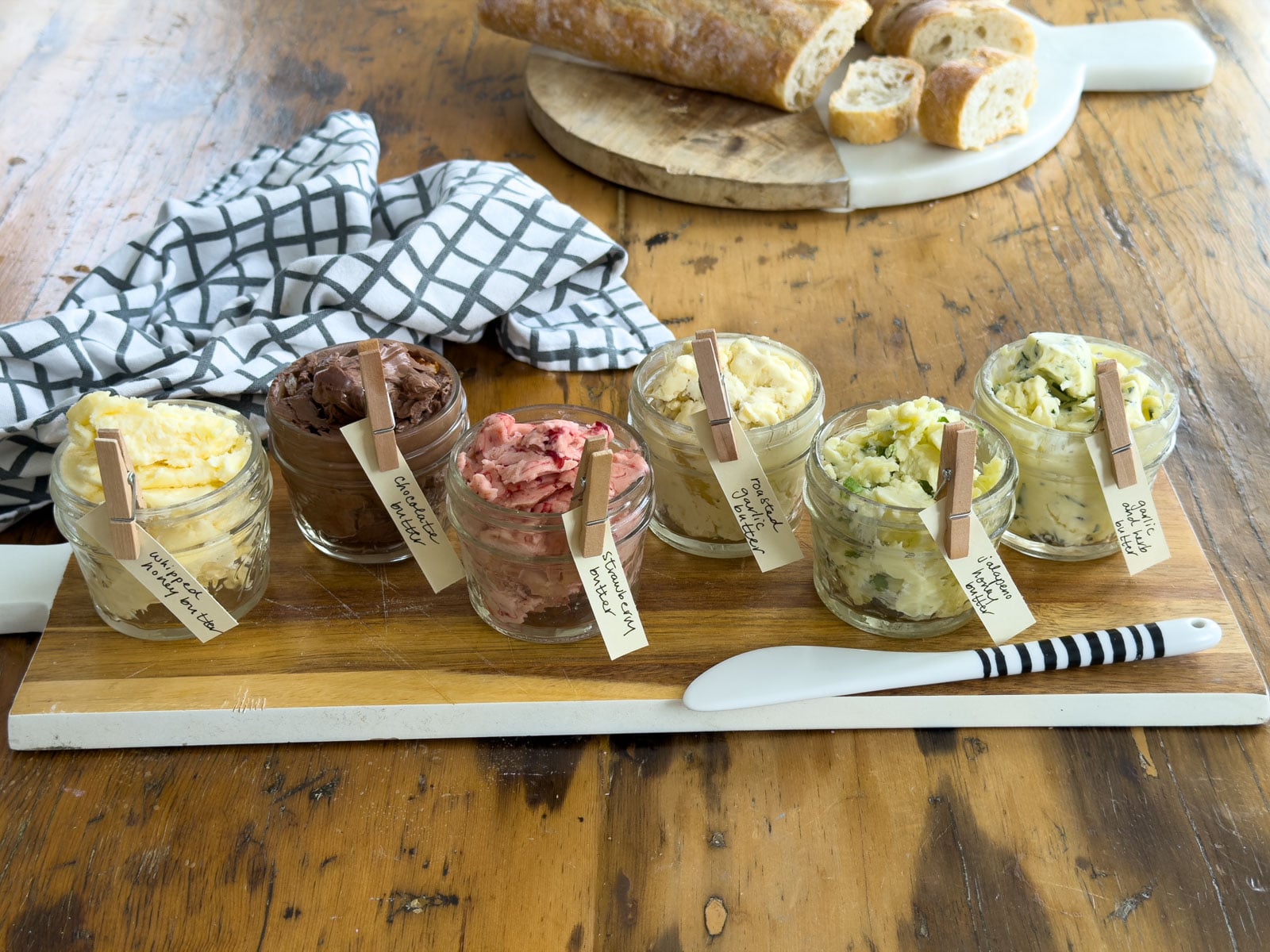 Overhead view of six compound butter jars with handwritten clothespin labels arranged on a round marble and wood board alongside a rectangular board with a sliced baguette on a wooden table.