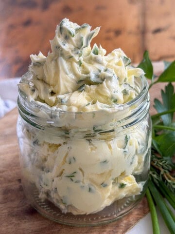 Garlic herb compound butter piled high in a glass jar with fresh herb flecks throughout, with parsley, thyme, and chives alongside.
