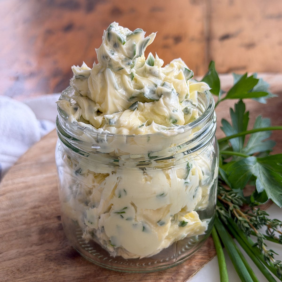 Garlic herb compound butter piled high in a glass jar with fresh herb flecks throughout, with parsley, thyme, and chives alongside.
