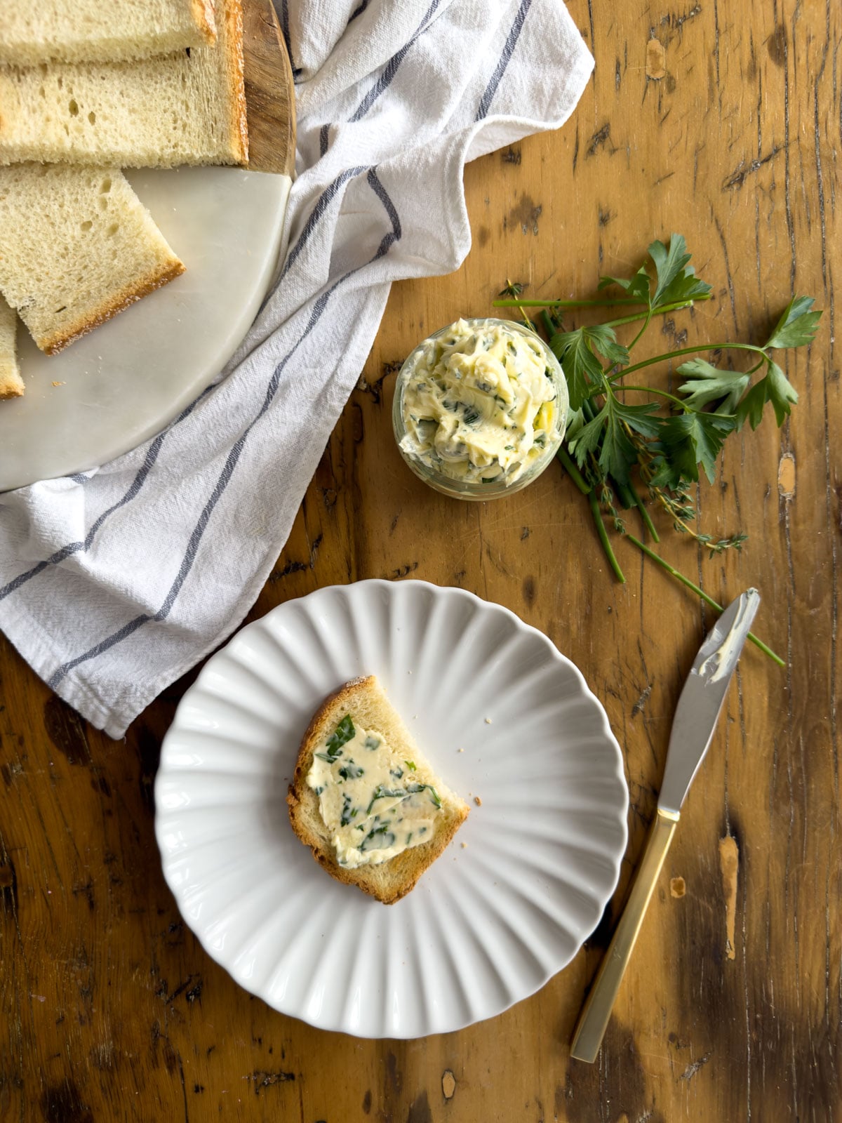 Overhead view of garlic herb compound butter spread on toast on a white scalloped plate with fresh herbs and bread on a wooden table.