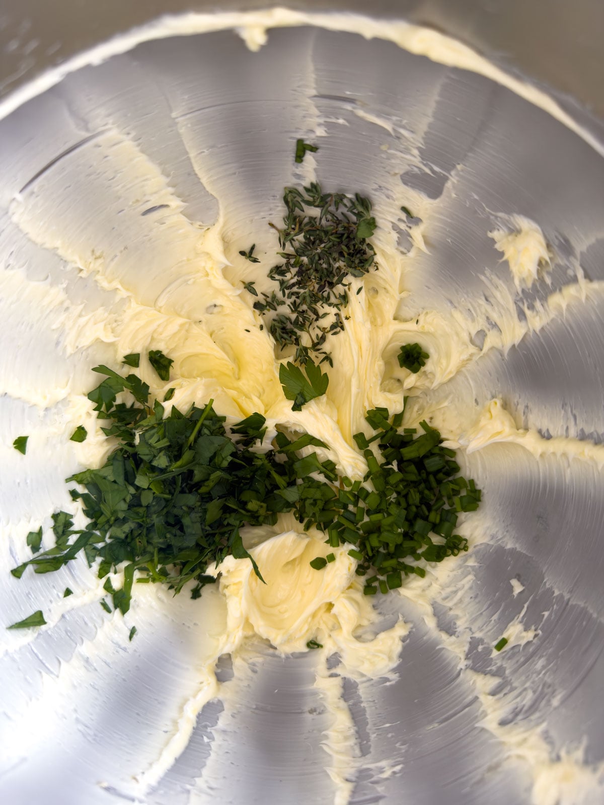 Chopped fresh parsley, chives, and thyme added to whipped butter in a stand mixer bowl before mixing.