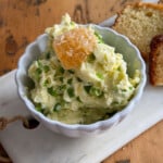 Jalapeno honey butter in a white scalloped bowl topped with a piece of honeycomb, served on a marble board with sliced bread in the background.
