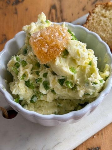 Jalapeno honey butter in a white scalloped bowl topped with a piece of honeycomb, served on a marble board with sliced bread in the background.