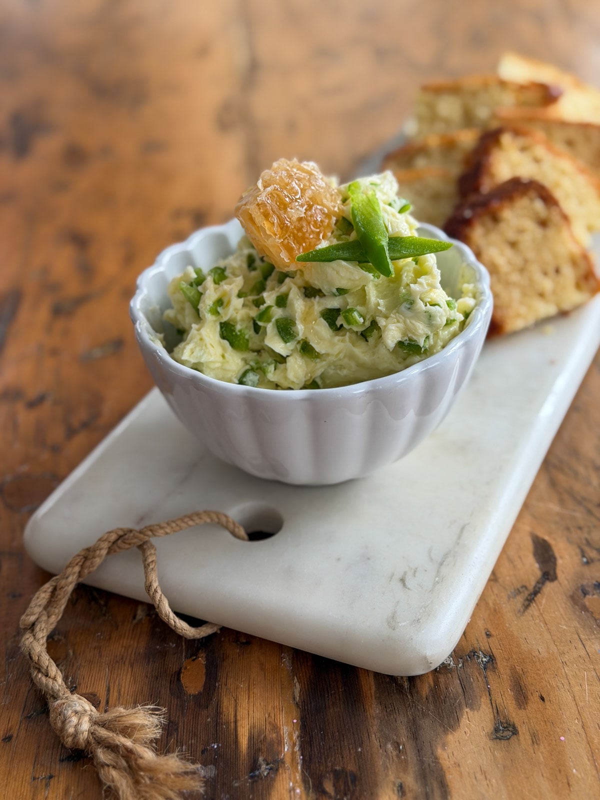Finished jalapeno honey butter in a white scalloped ramekin garnished with honeycomb and a jalapeno slice, served on a marble board with sliced bread, ready to refrigerate or serve.