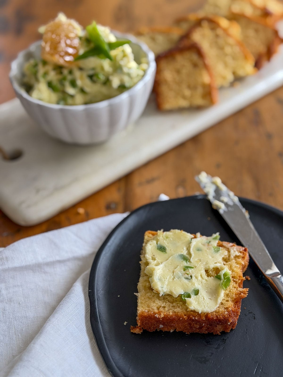 Jalapeno honey butter in a white scalloped bowl on a marble board with sliced bread, and a piece of cornbread spread with jalapeno honey butter on a black plate in the foreground.