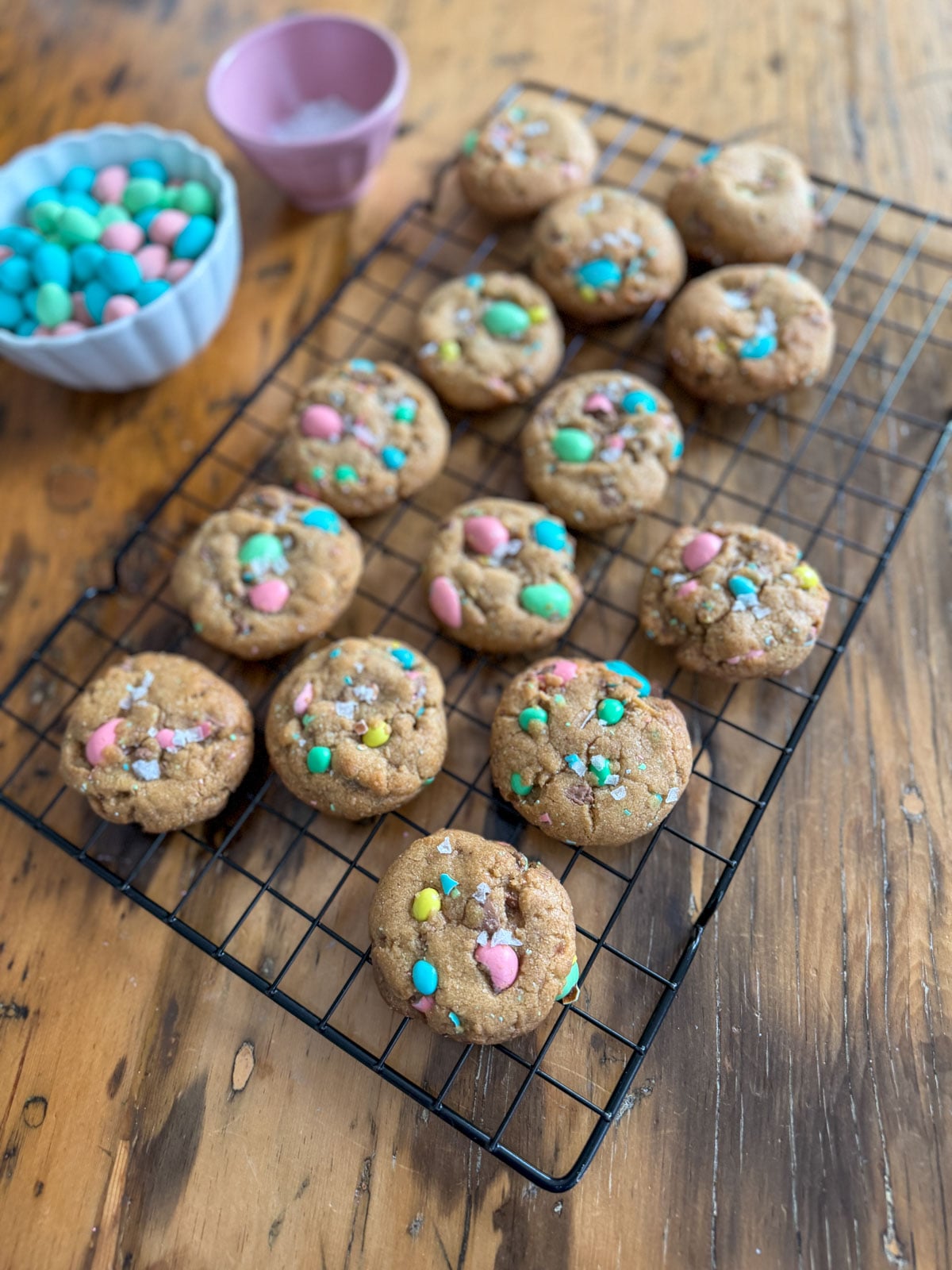 Brown butter Cadbury mini egg cookies on a wire rack with flaky salt, a pink mug of tea, and a bowl of mini eggs.