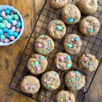 Cadbury mini egg cookies with flaky sea salt cooling on a wire rack with a bowl of pastel mini eggs alongside.