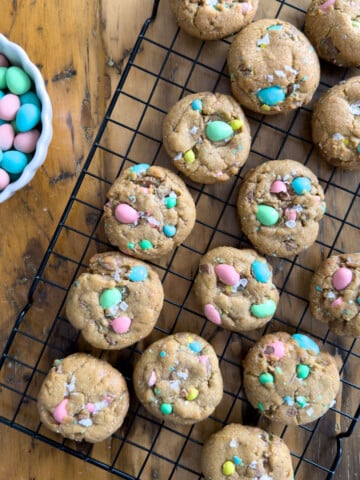 Cadbury mini egg cookies with flaky sea salt cooling on a wire rack with a bowl of pastel mini eggs alongside.