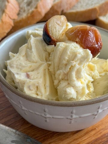 Whipped roasted garlic butter topped with golden caramelized garlic cloves in a white embossed ceramic bowl with sliced bread in the background.