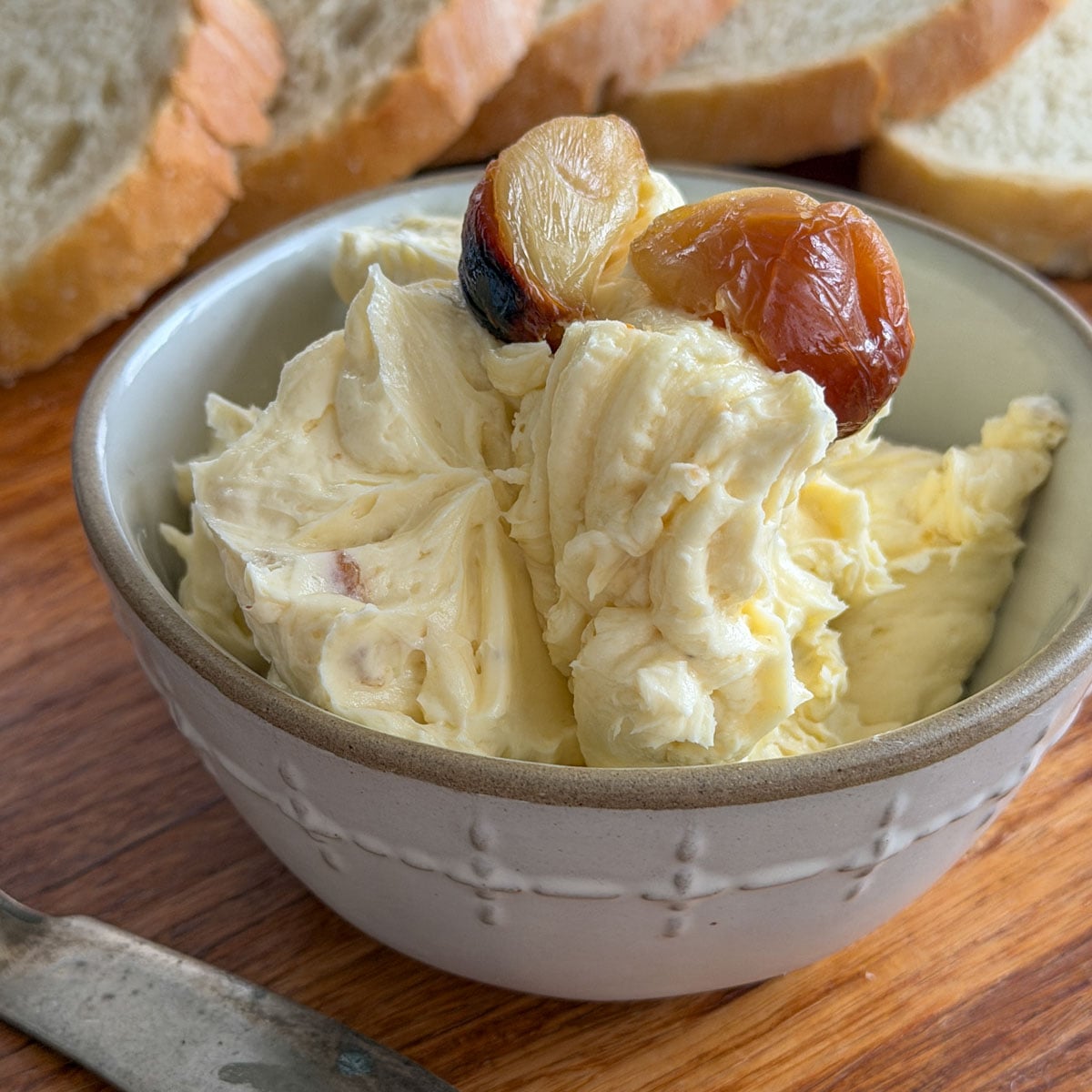 Whipped roasted garlic butter topped with golden caramelized garlic cloves in a white embossed ceramic bowl with sliced bread in the background.