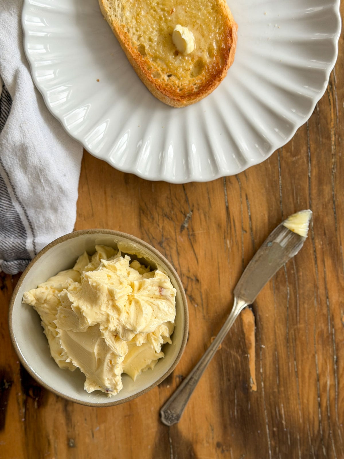 Overhead view of whipped roasted garlic butter in a ceramic bowl with a silver butter knife and buttered toast on a white scalloped plate on a wooden table.