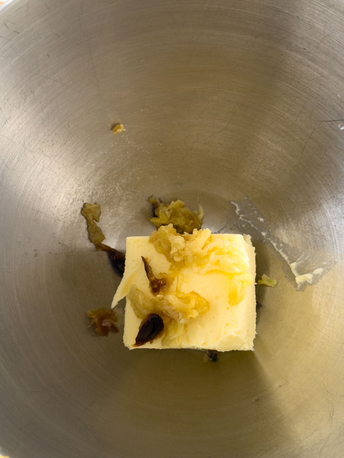 Overhead view of a block of softened butter with squeezed roasted garlic cloves on top in a stand mixer bowl.