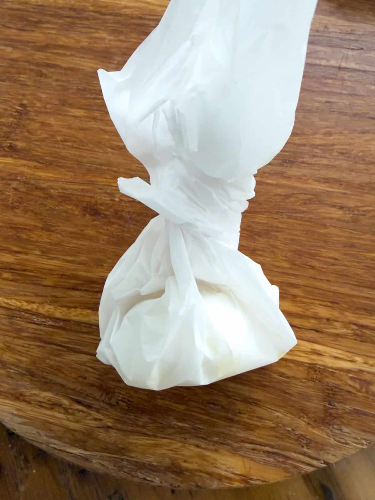 Garlic head wrapped in parchment paper and twisted into a bundle ready for roasting on a wooden cutting board.