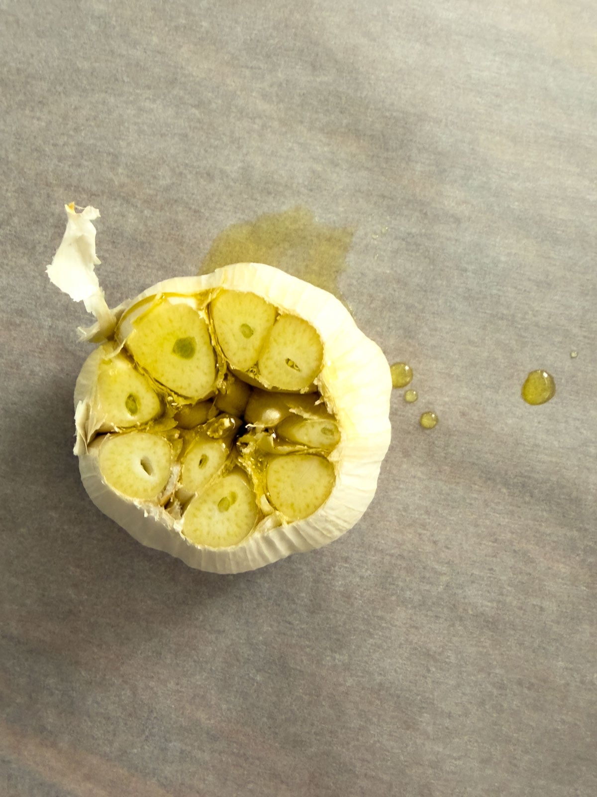 Overhead view of a garlic head with the top sliced off and olive oil drizzled over the exposed cloves on parchment paper.