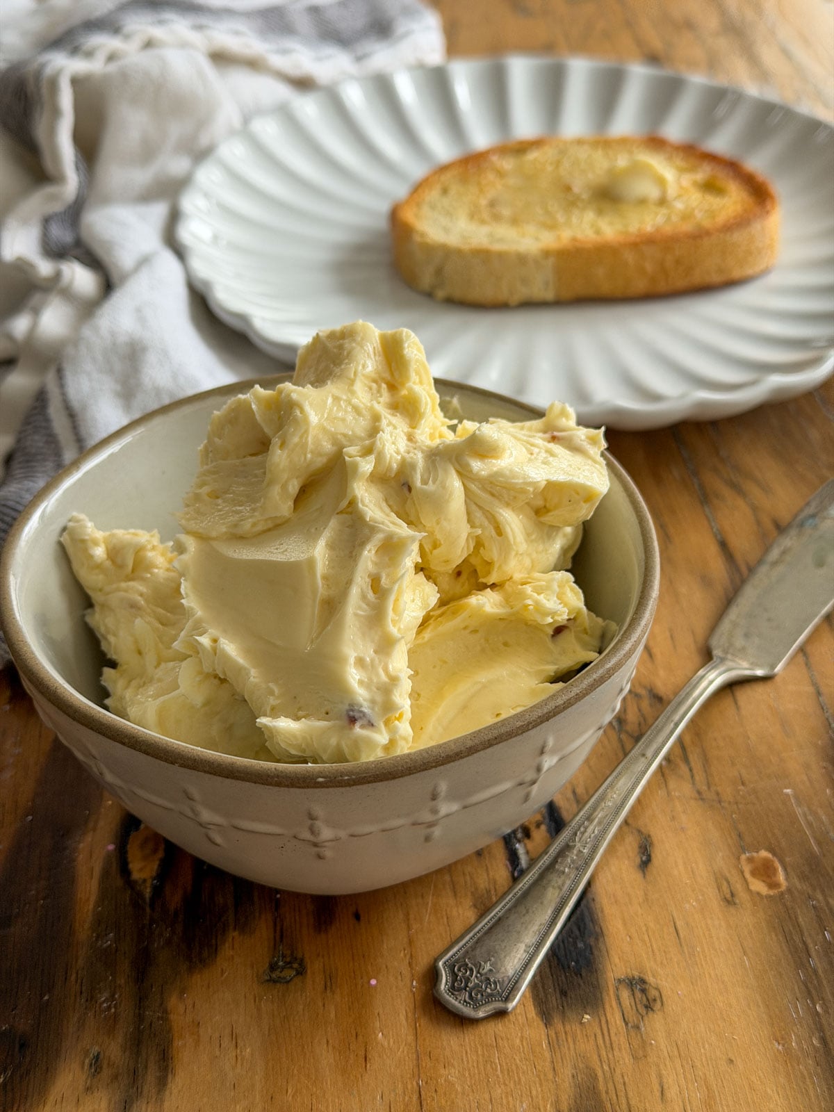 Close-up of freshly whipped roasted garlic butter in a white embossed ceramic bowl with toast on a scalloped plate in the background.