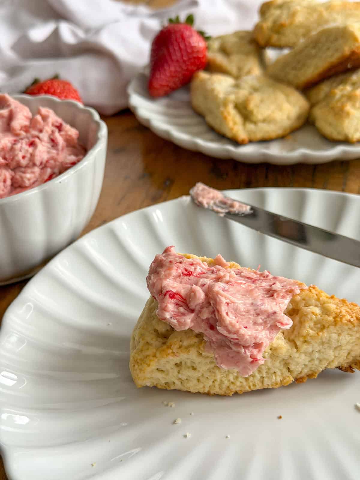  Triangle scone spread with pink whipped strawberry butter on a white scalloped plate, with a butter knife resting on the plate, a bowl of strawberry butter and a plate of scones in the background.