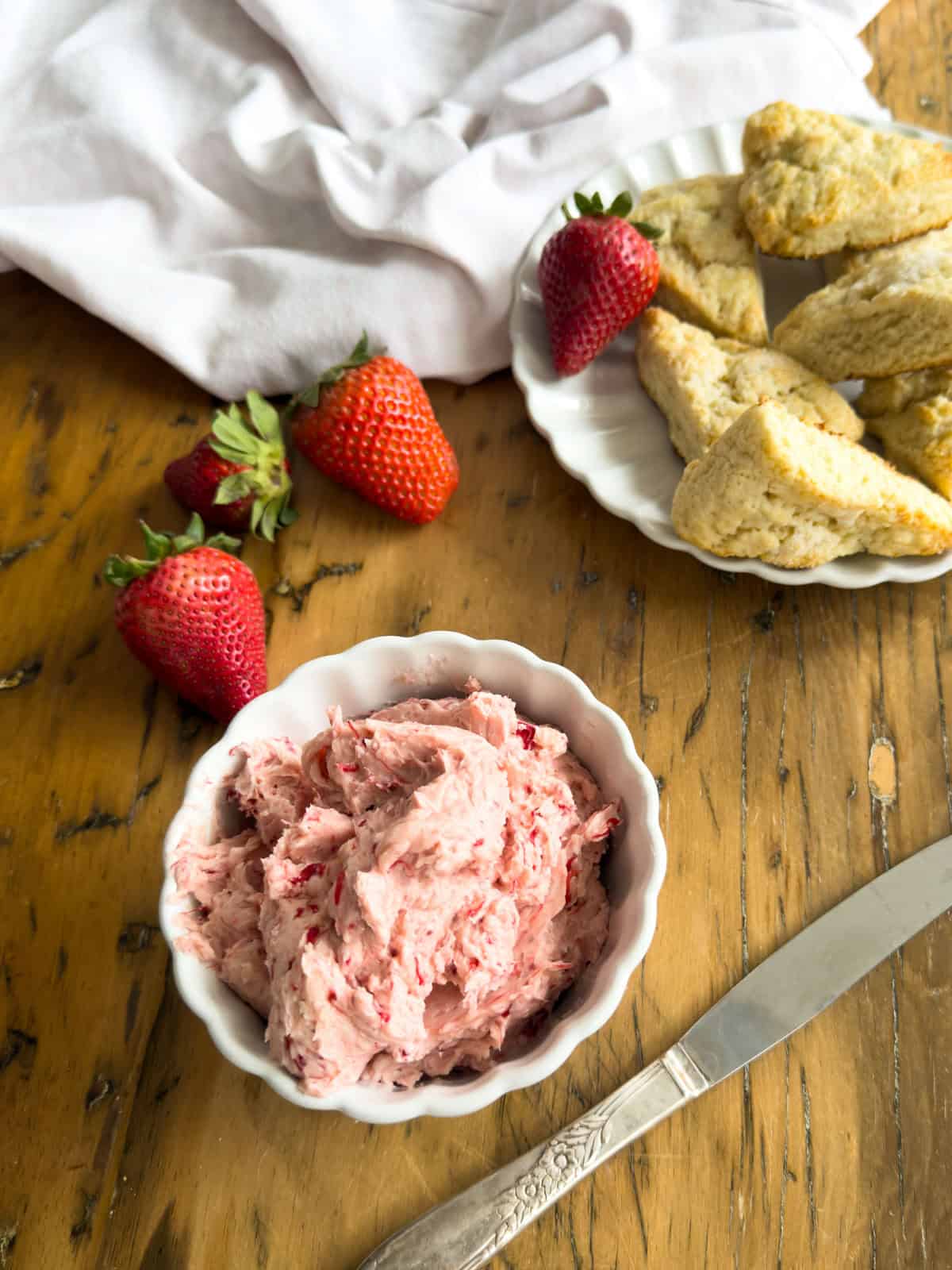 Overhead view of pink whipped strawberry butter in a white scalloped bowl surrounded by fresh strawberries, a butter knife, and a plate of triangle scones on a wood table.