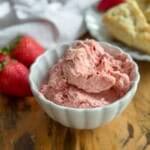 Whipped strawberry butter in a white scalloped bowl with two fresh strawberries to the left and golden scones on a white plate in the background.