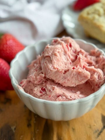 Whipped strawberry butter in a white scalloped bowl with two fresh strawberries to the left and golden scones on a white plate in the background.