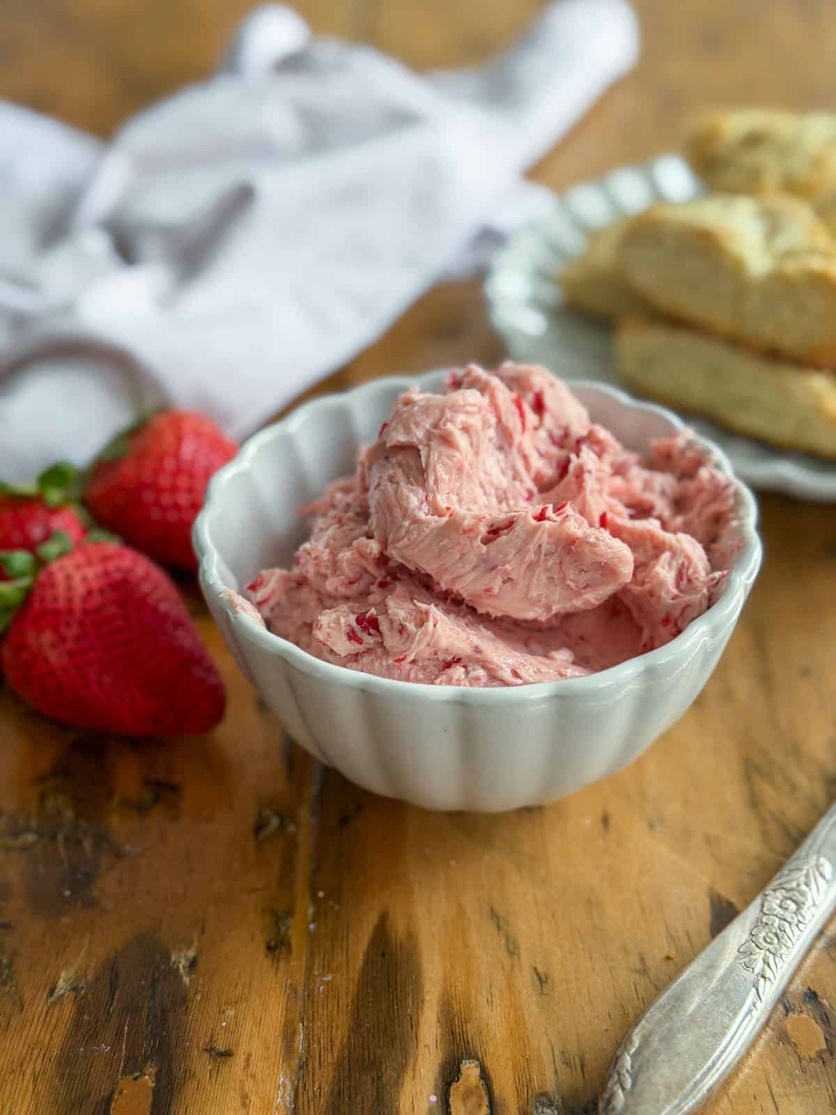 Whipped strawberry butter mounded in a white scalloped bowl with two fresh strawberries to the left and scone pieces softly visible in the background.