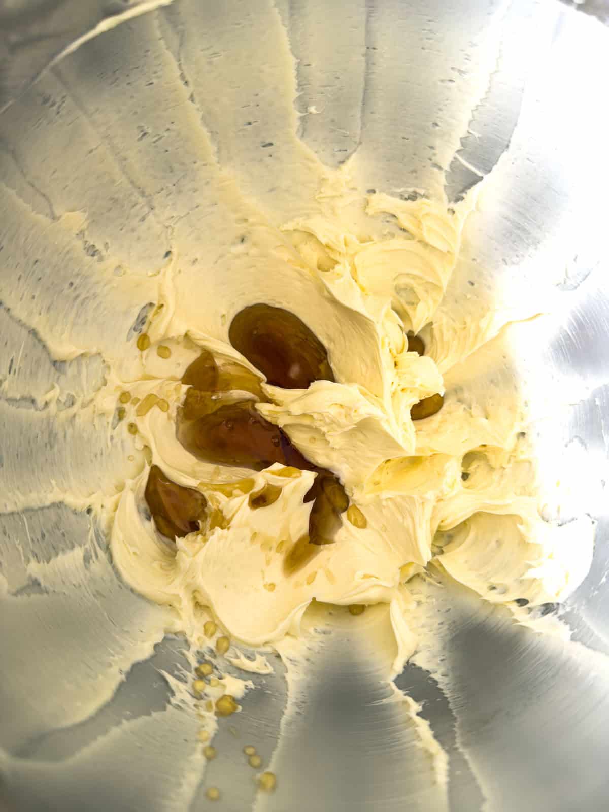 Pale yellow whipped butter in a stainless steel stand mixer bowl with amber maple syrup pooled in the center, ready to be mixed in.