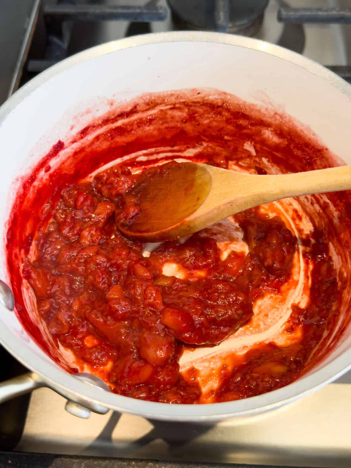 Overhead view of strawberry butter ingredients on a wood table: a glass pitcher of maple syrup, a white scalloped bowl of frozen strawberries, and a white scalloped bowl with a block of butter, each labeled.