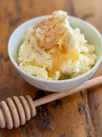 Whipped honey butter in a white ceramic bowl topped with a piece of golden honeycomb and a wooden honey dipper on a rustic wooden table.