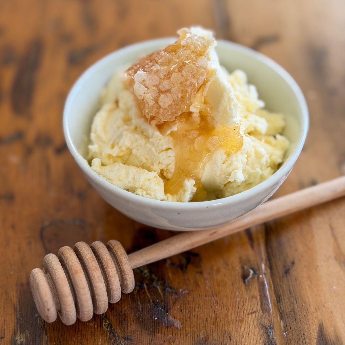 Whipped honey butter in a white ceramic bowl topped with a piece of golden honeycomb and a wooden honey dipper on a rustic wooden table.