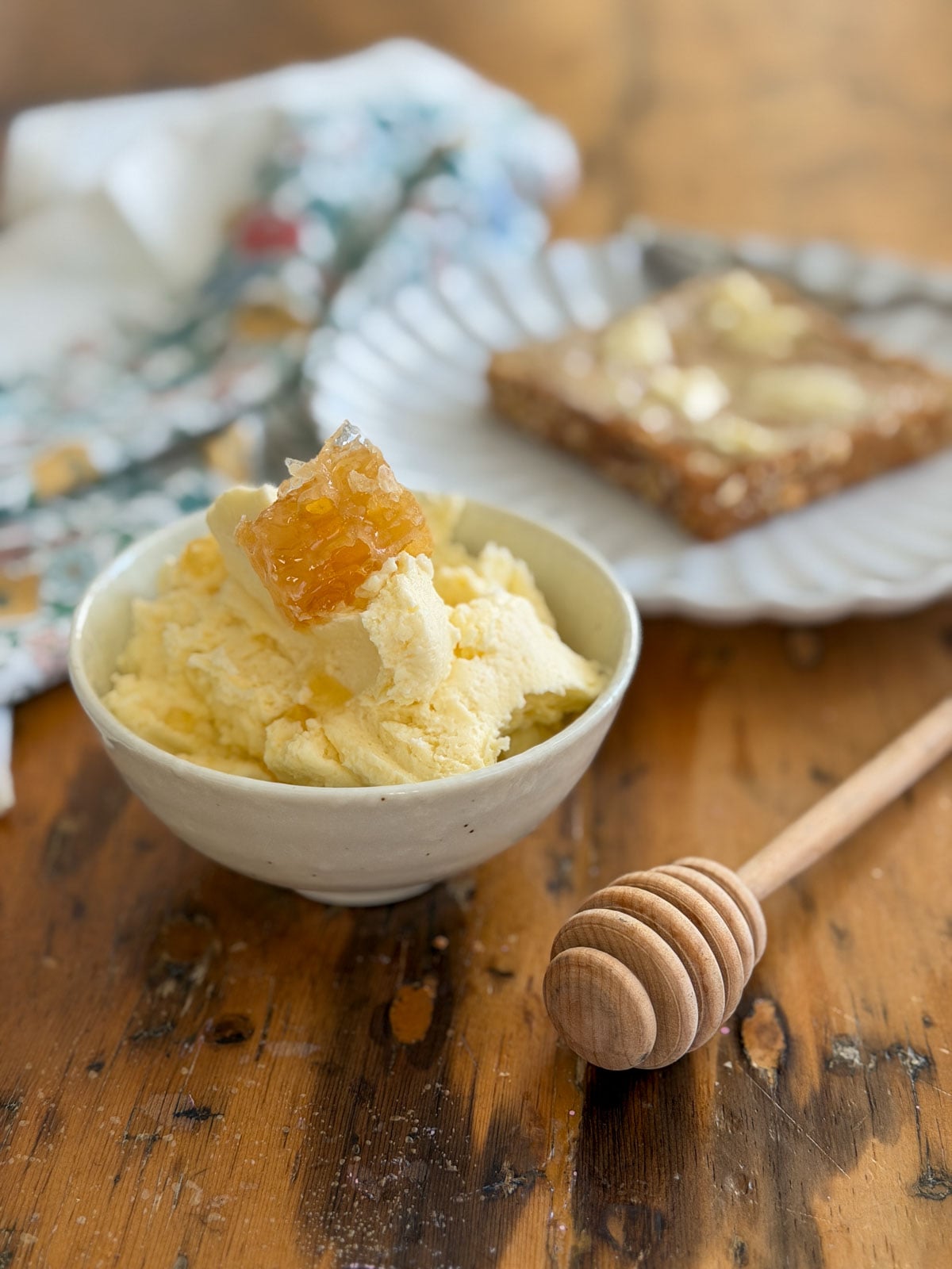 Finished whipped honey butter in a white ceramic bowl topped with a golden honeycomb piece and honey drizzle, with a wooden honey dipper on a rustic wooden table.