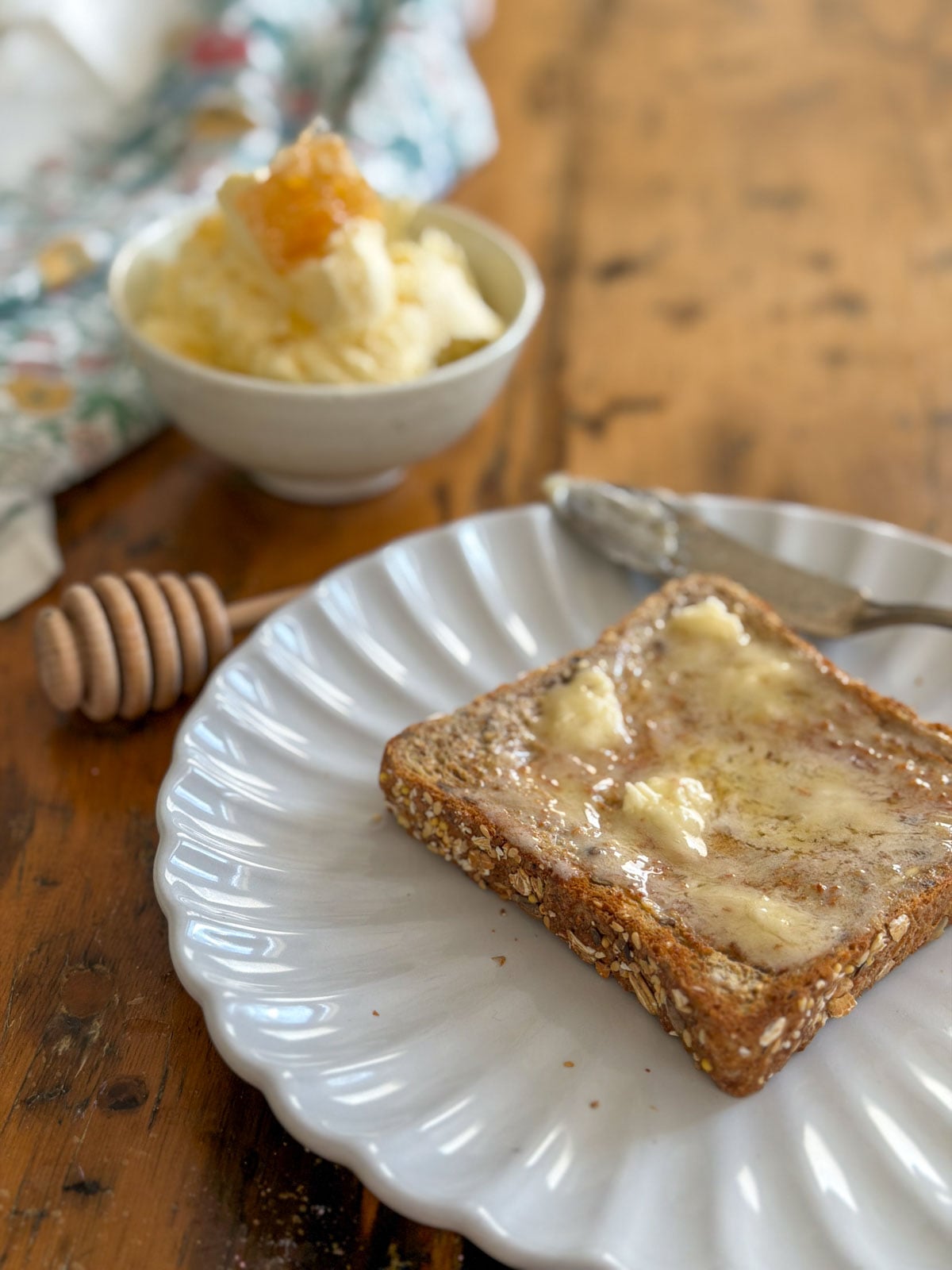 Whipped honey butter melting on a slice of multigrain toast on a white scalloped plate with a bowl of honey butter topped with honeycomb and a wooden honey dipper visible in the background.