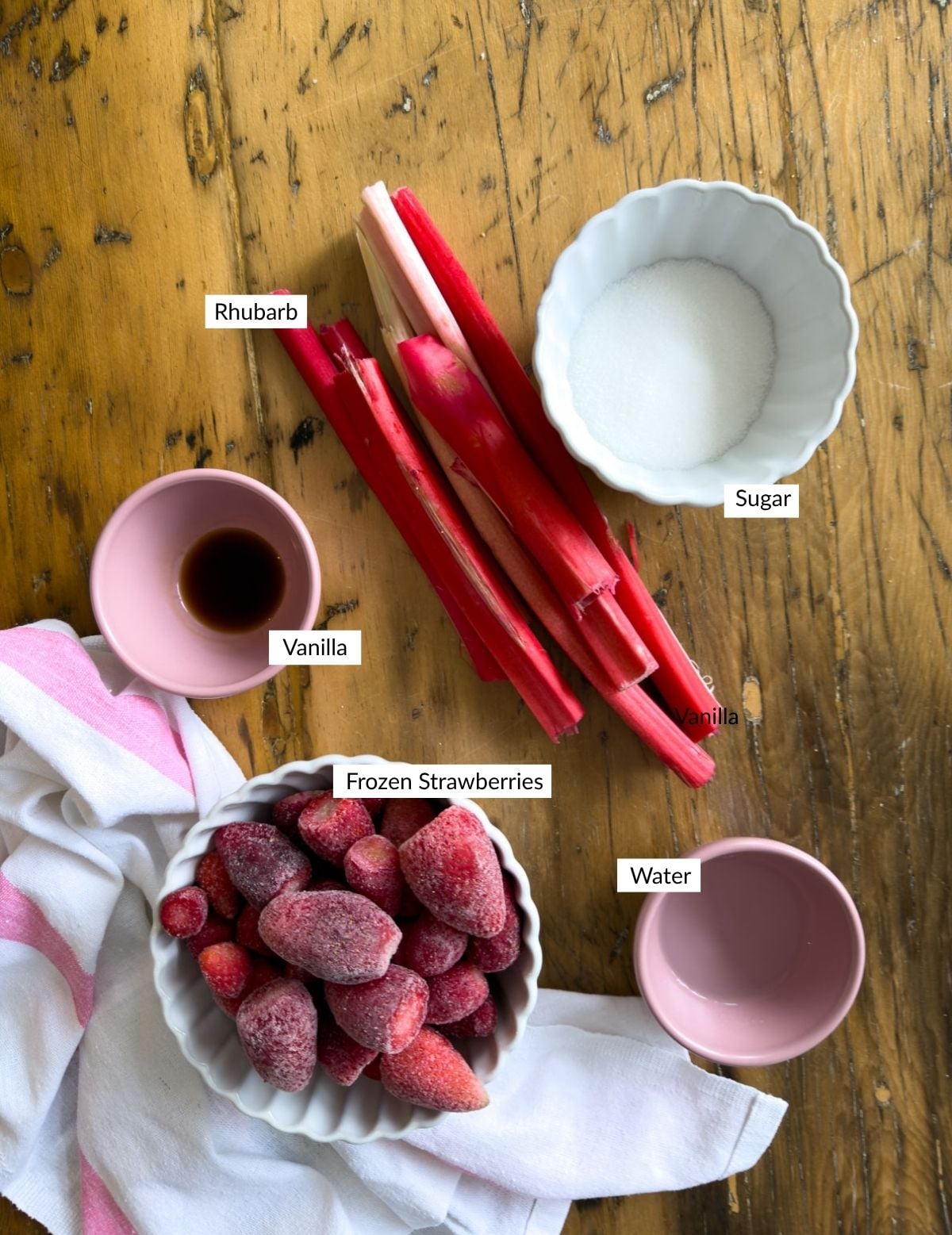 Ingredients for strawberry rhubarb compote arranged on a wooden surface including a bowl of frozen strawberries, fresh rhubarb stalks, a bowl of white granulated sugar, a small bowl of lemon juice, and a pink cup of vanilla extract.