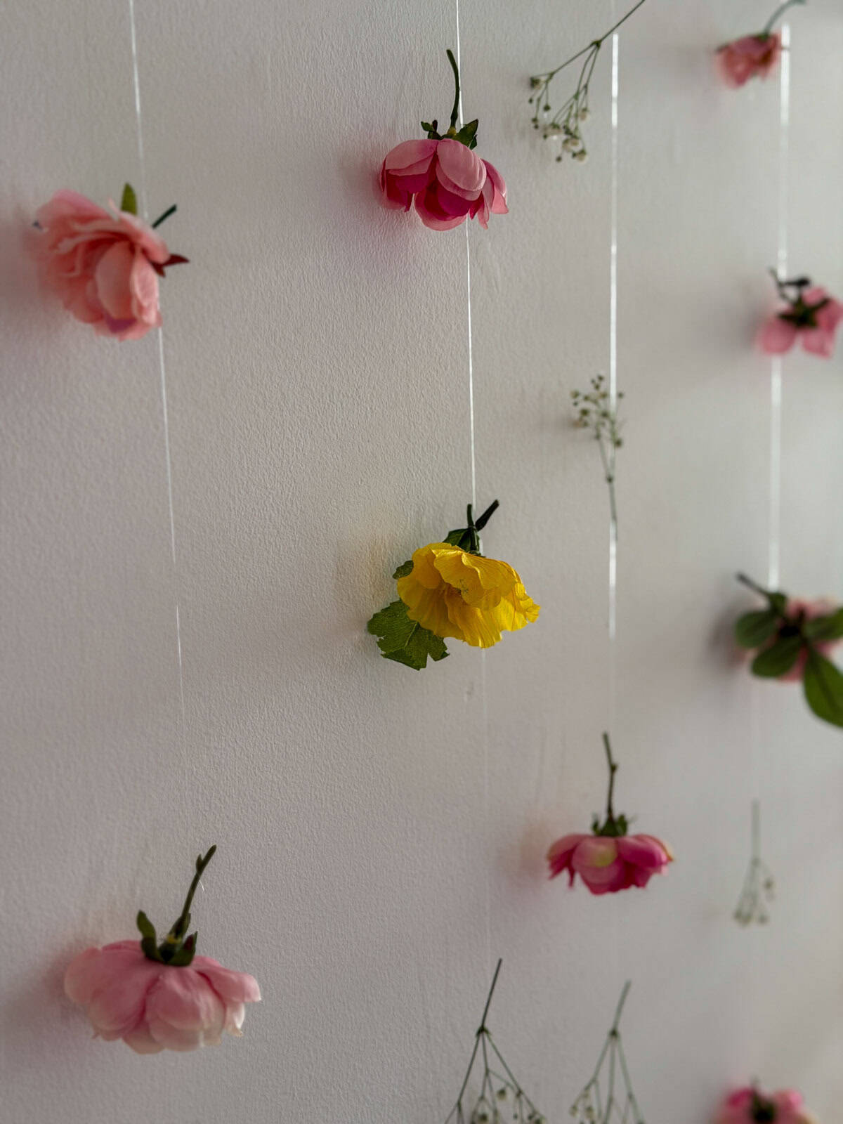 Pink and yellow faux flowers and baby's breath suspended on clear strings against a white wall in a cascading flower curtain installation.