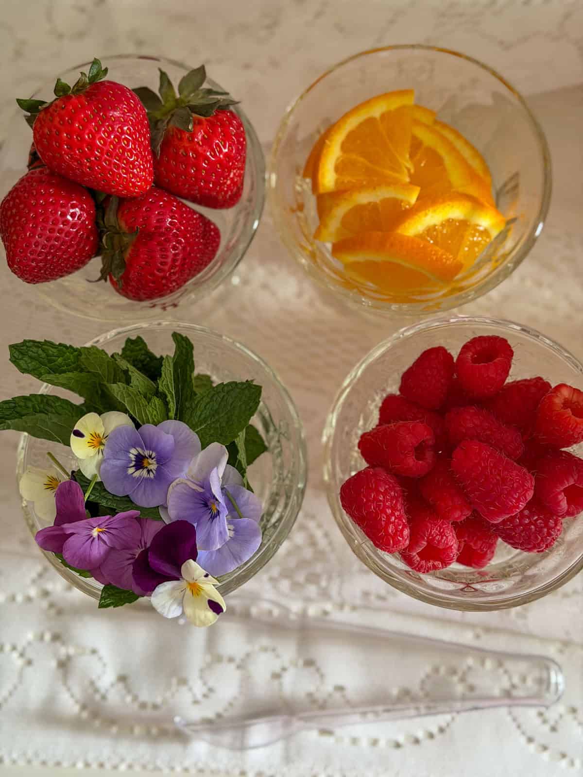 Four glass bowls of mimosa bar garnishes including fresh strawberries, orange slices, edible pansies with mint and raspberries on white lace tablecloth.