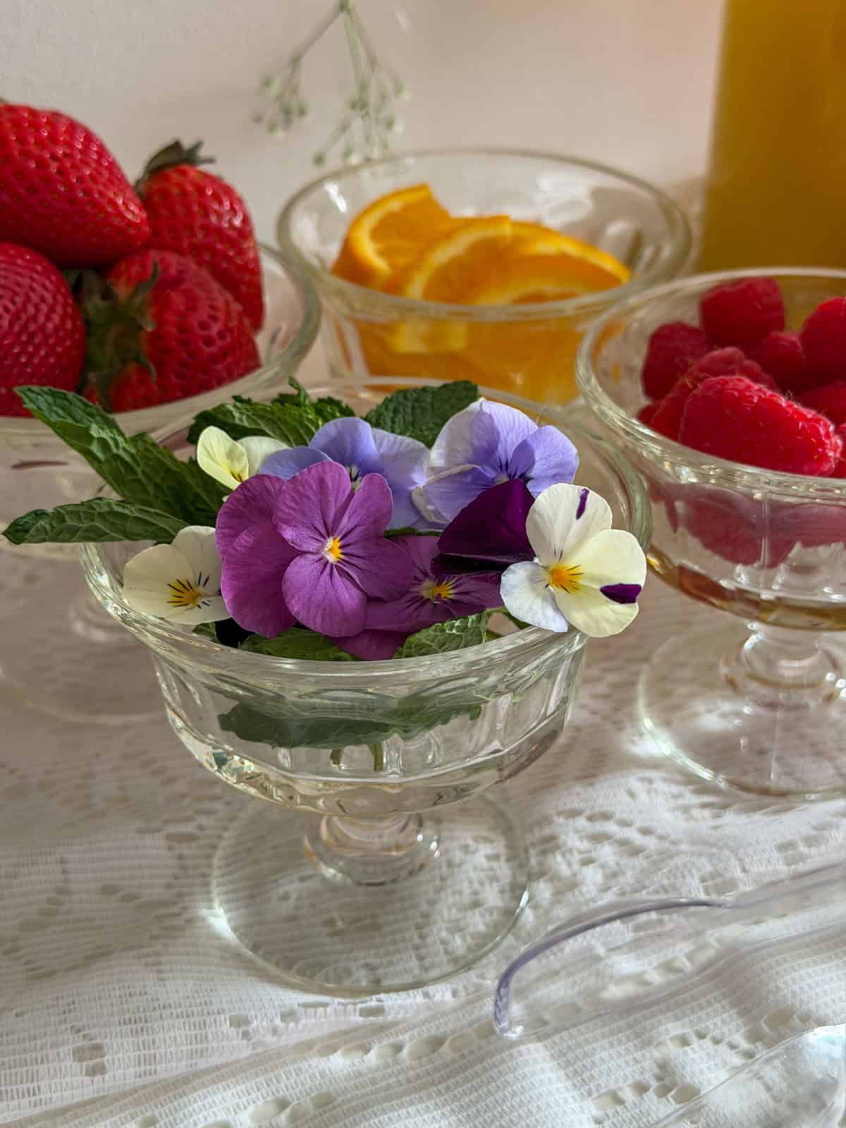 Glass pedestal bowl filled with purple and white edible pansy flowers and fresh mint leaves with strawberries and raspberries visible behind.